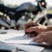 Mãos assinando um contrato com a moto ao fundo, ideia de conferir se a associação de proteção veicular é confiável