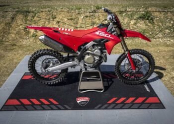 Ducati Desmo450 MX vermelha sobre tapete da Ducati Corse, com fundo de pista de terra