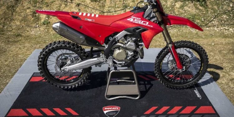 Ducati Desmo450 MX vermelha sobre tapete da Ducati Corse, com fundo de pista de terra