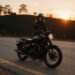Mulher motociclista sorri ao lado da moto numa estrada ao pôr do sol, para frases de moto feminina