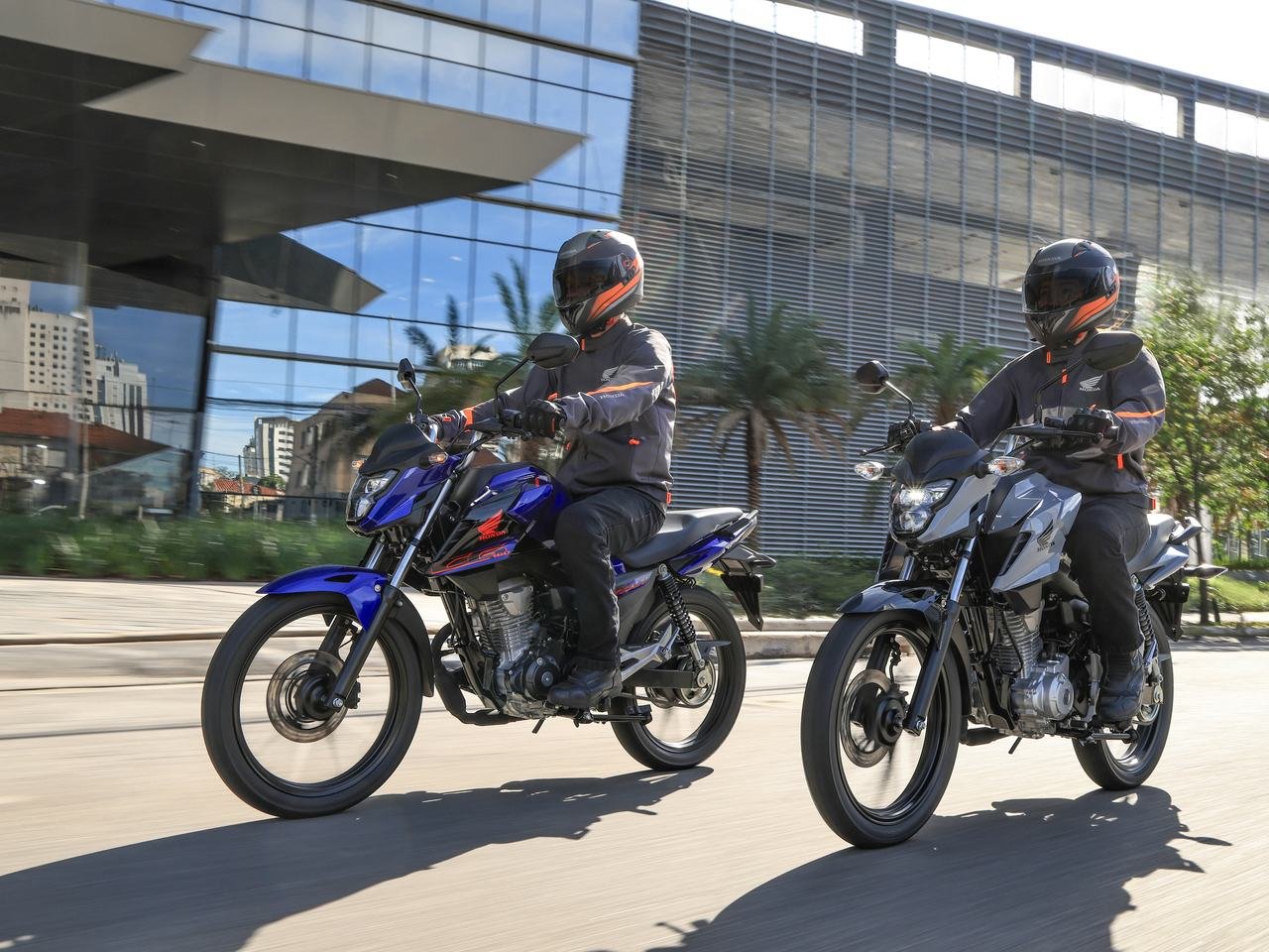Duas motos Honda 2027, uma azul e uma cinza, rodando em frente a prédios