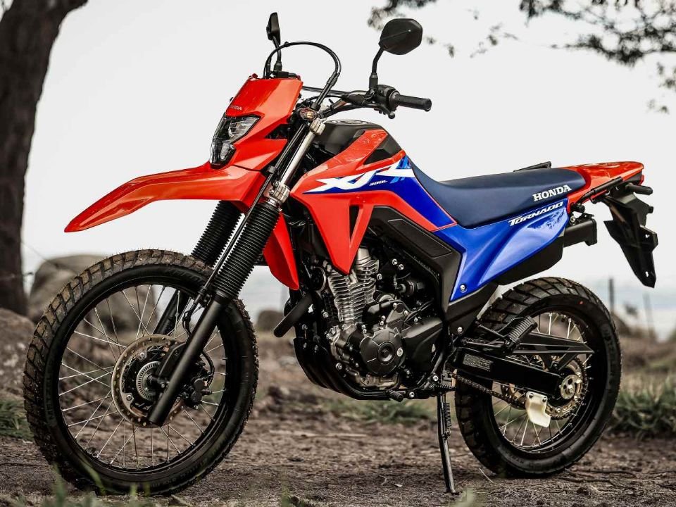 Honda XR300L Tornado 2027 vermelha e azul parada, vista lateral com rodas raiadas