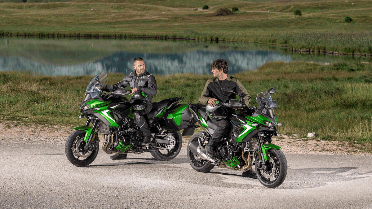 Dois pilotos ao lado de duas Kawasaki Versys 900 2027 verdes com malas laterais, em paisagem com lago