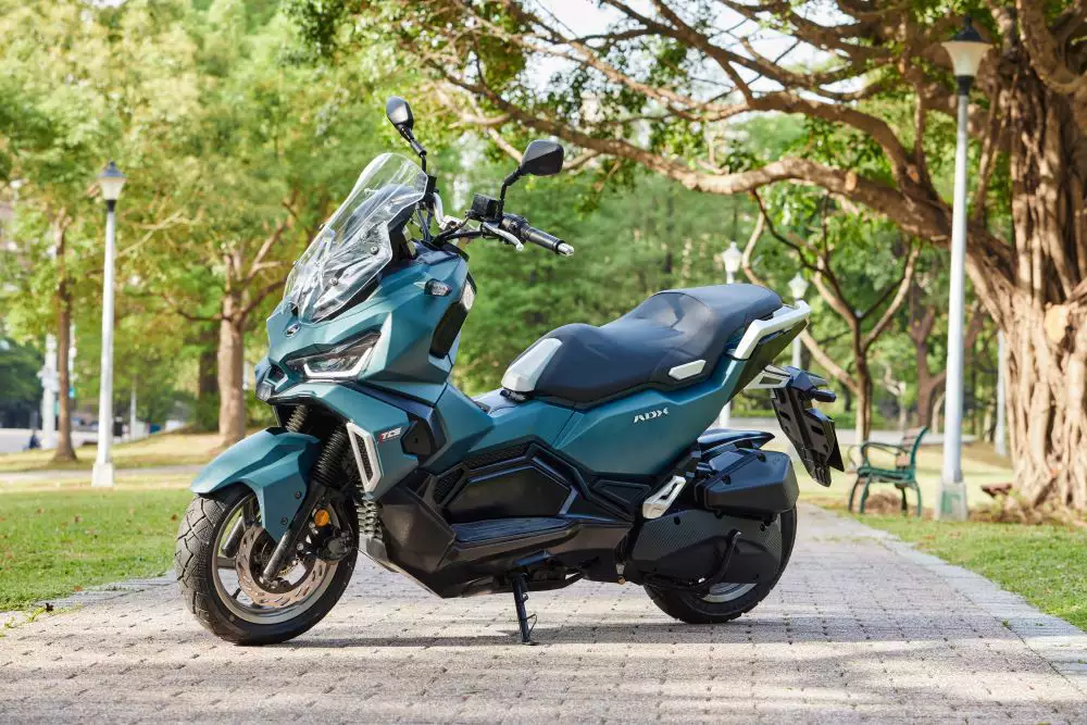 Scooter SYM ADX em tom azul-esverdeado parada em calçada de parque, modelo da linha com revisão a preço fixo