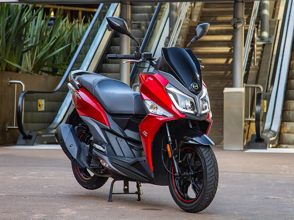 Scooter SYM vermelha com rodas pretas e friso vermelho, parada diante de escada rolante